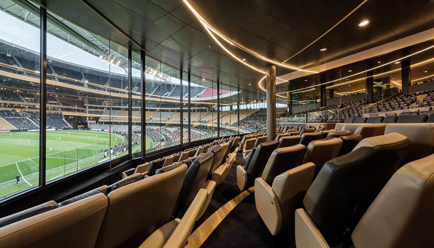 Stadium luxury suite with rows of leather recliner seats overlooking a green pitch and crowd, warm lighting